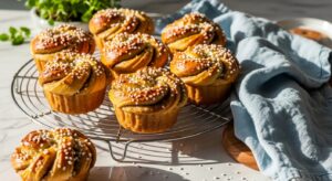 A beautifully arranged shot of several golden brown, pearl sugar-topped cardamom buns, baked in muffin-like shapes with visible swirls, resting on a silver wire cooling rack. A light blue linen cloth is artfully draped nearby. The scene is set on a marble countertop with subtle wood accents, bathed in natural morning light, creating soft shadows and a warm, inviting atmosphere. Fresh herbs are visible in the soft background. No hands.