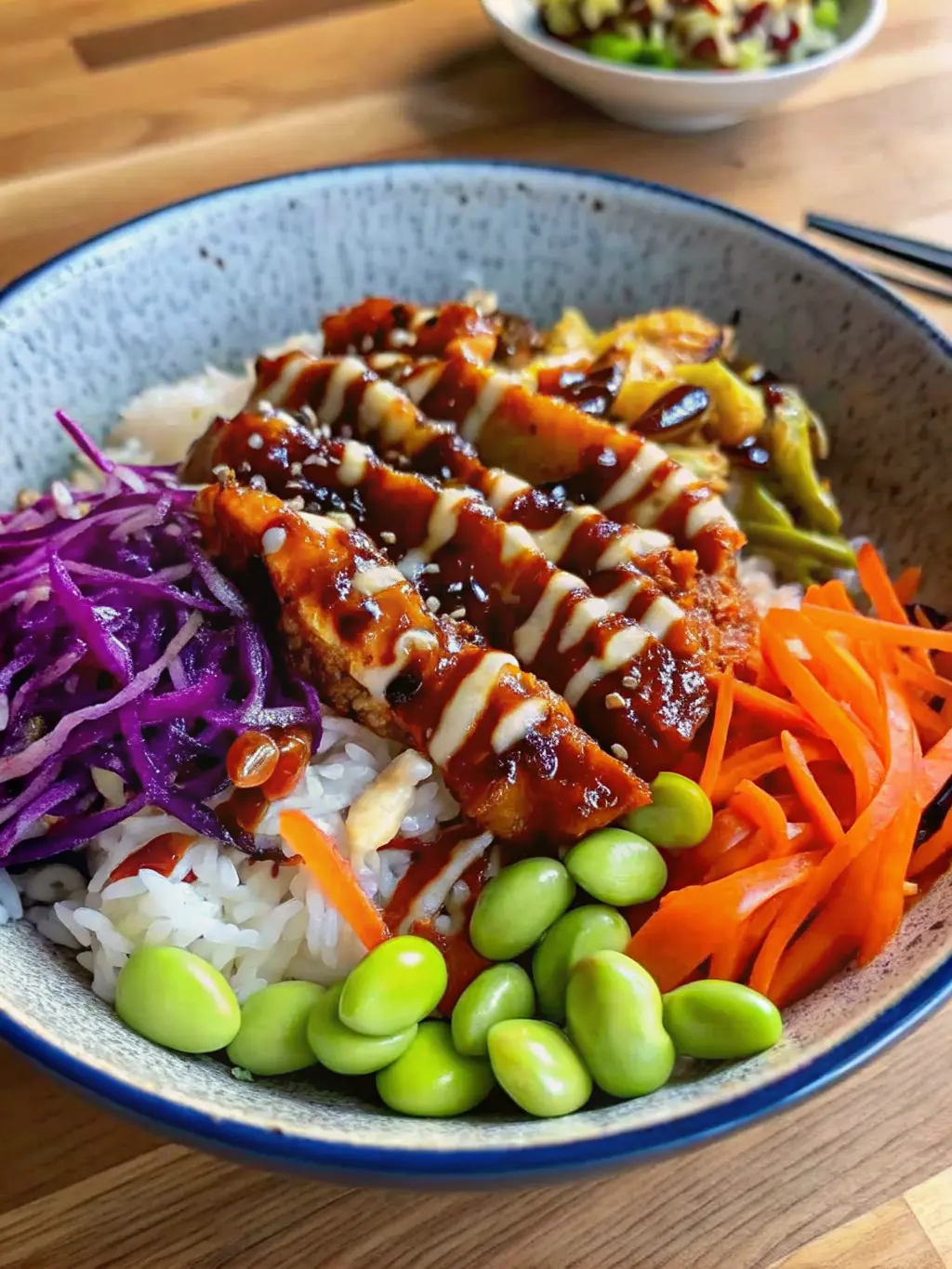 A beautifully composed serving shot of a Teriyaki Chicken Bowl (3:4 ratio), showcasing the distinct textures and colors. The focus is on the tender chicken glazed with teriyaki sauce, the crispness of the shredded carrots and red cabbage, the plump edamame, and the refreshing pickled cucumber, all over fluffy white rice in a blue ceramic bowl. A drizzle of creamy white sauce and sesame seeds add visual interest. The bowl rests on a wooden surface, with soft, warm light emphasizing the textures. NO HANDS OR PEOPLE.