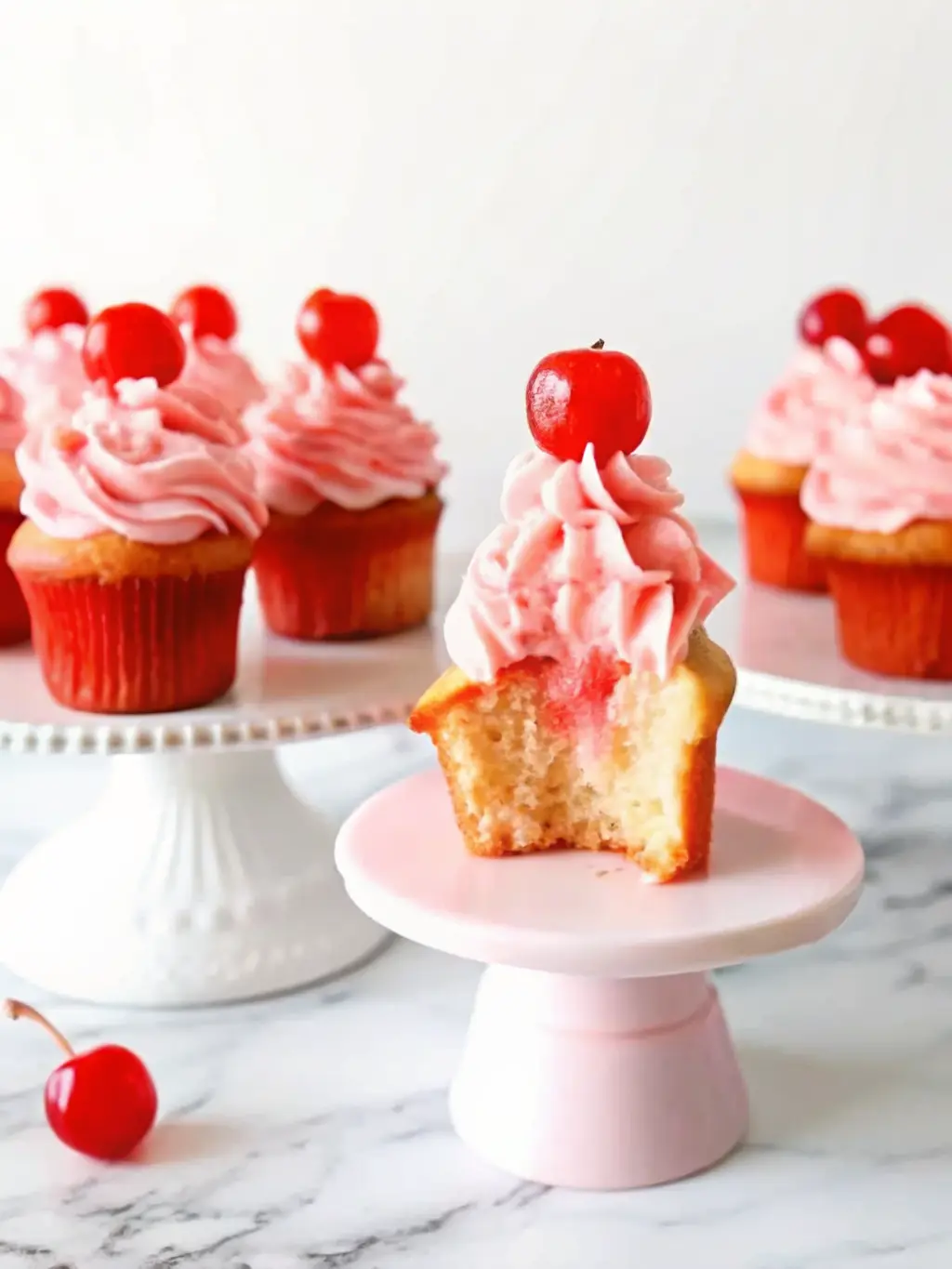 Close-up shot of the cupcake batter, visibly pink from cherry juice, being gently folded with chopped maraschino cherries in a minimalist white ceramic bowl, sitting on a white marble countertop. A wooden spoon is resting in the bowl. Soft morning light and warm tones highlight the texture. 3:4 ratio. NO HANDS OR PEOPLE.