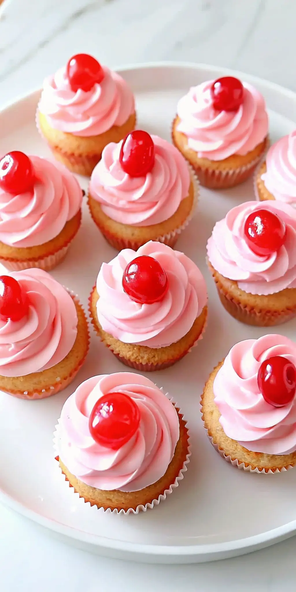 A close-up, artful shot of a frosted Cherry Chip Cupcake, displaying the elegant pink swirled buttercream frosting and the vibrant red maraschino cherry on top, showcasing the texture. The cupcake is on a white plate, positioned on a white marble countertop in natural morning light. Soft shadows, warm tones, clean presentation. 3:4 ratio. NO HANDS OR PEOPLE.