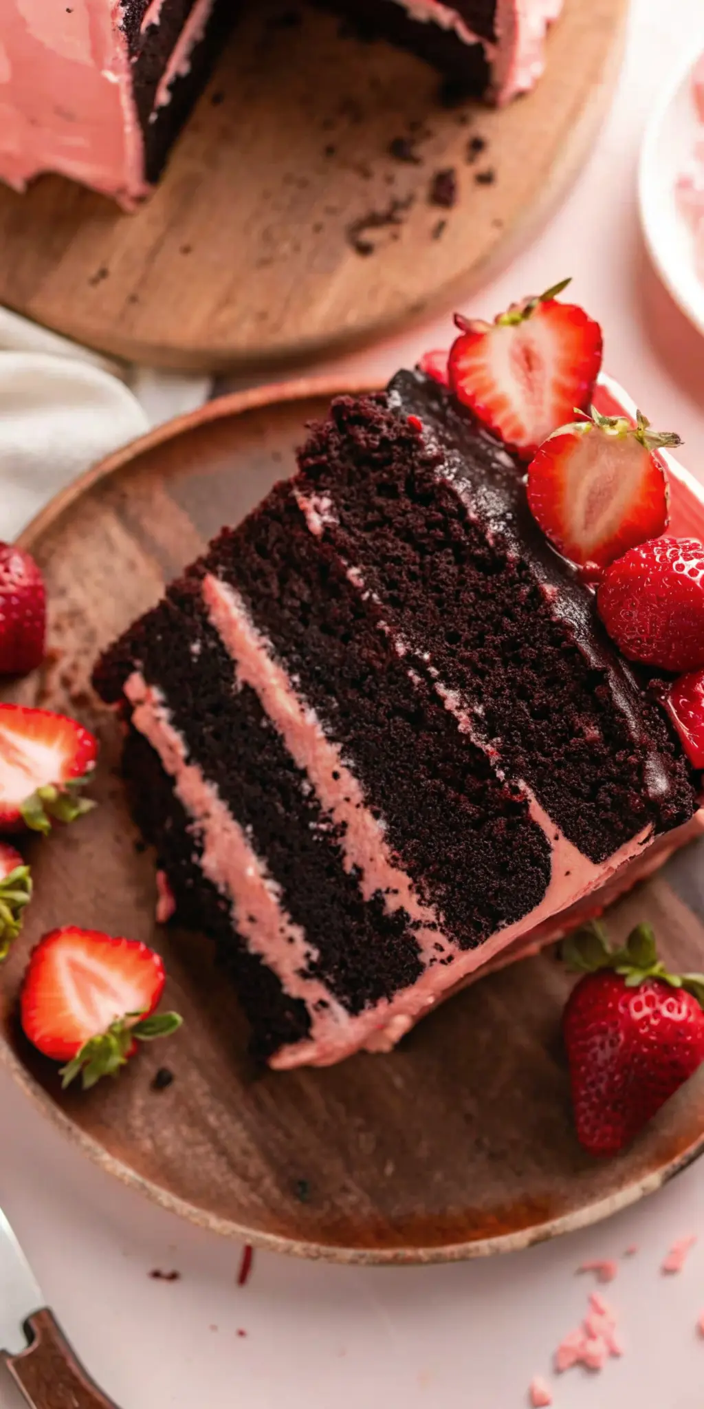 A close-up, slightly elevated shot of a perfectly cut slice of Chocolate Covered Strawberry Cake, revealing the moist chocolate cake texture and the smooth, airy pink strawberry buttercream layers. A rich chocolate ganache drip is visible, and fresh strawberries are artfully placed on the side. The cake slice is on a minimalist white plate, on the familiar wooden cutting board on a marble countertop, with warm, soft morning light and soft shadows, emphasizing the deliciousness. NO HANDS. (3:4 ratio)