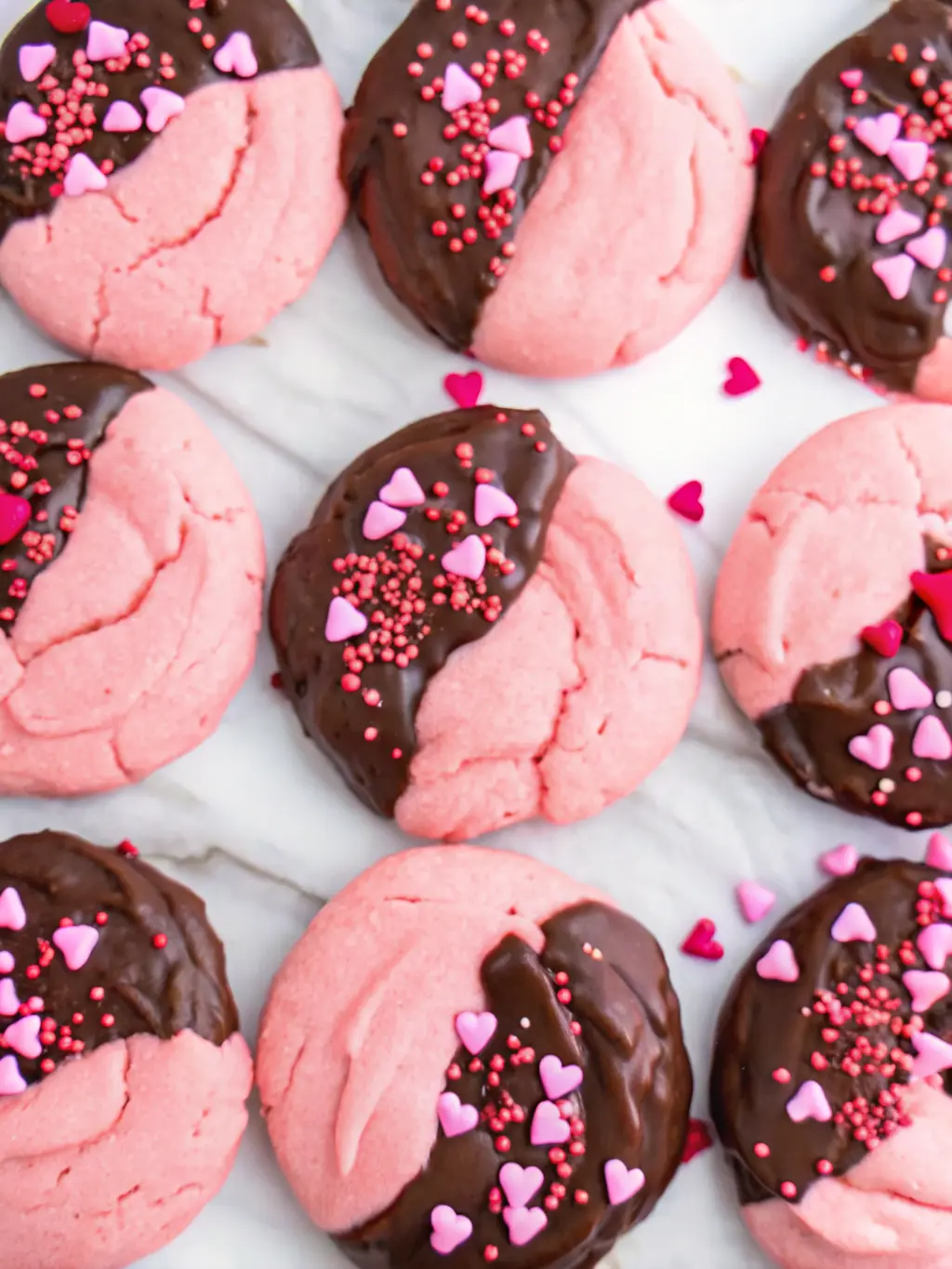 An overhead shot of key ingredients for Chocolate Covered Strawberry Cookies laid out on a wooden cutting board on a marble countertop: a bowl of vibrant pink cookie dough, a bar of dark chocolate, and a small ceramic dish filled with red, pink, and white heart sprinkles. Fresh herbs in the background. Natural morning light from an east window, soft shadows, warm tones, clean and tidy presentation. NO HANDS OR PEOPLE. (3:4 ratio)