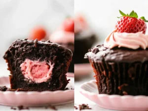 A close-up hero shot of a dark chocolate cupcake, cut in half to reveal a perfectly formed, vibrant pink, heart-shaped strawberry cream filling. The cupcake is topped with glossy dark chocolate ganache and a single fresh, ripe red strawberry. Crumbs are scattered artfully around the base. The setting is a clean marble countertop with soft natural morning light from an east window, subtle warm tones, and soft shadows. A wooden cutting board is subtly visible in the background, along with a minimalist white plate holding other cupcakes, and fresh green herbs are softly blurred in the deep background. No hands or people. (4:3 ratio)