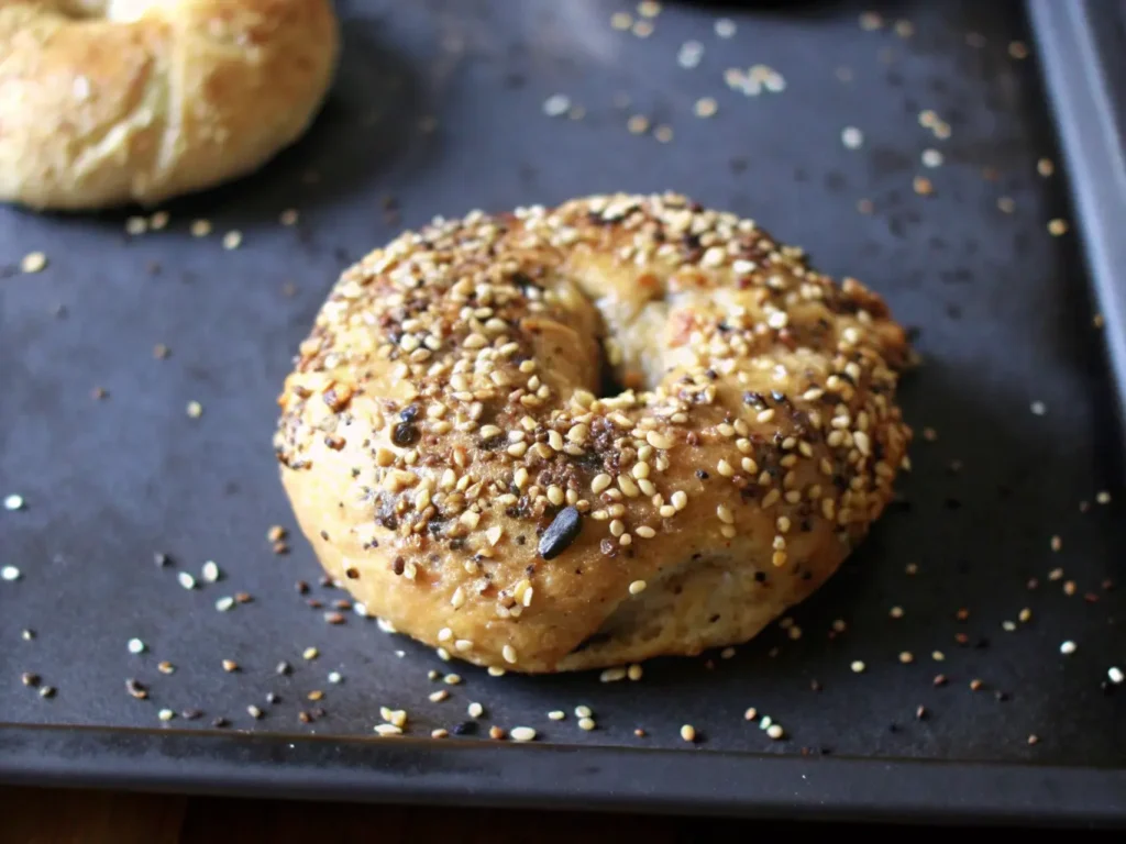 A single Greek Yogurt Bagel, broken in half, showcasing its chewy, golden-brown crust and soft interior. Generously topped with poppy seeds and sesame seeds. The bagel rests on a dark baking sheet, with scattered seeds and slight caramelized residue. Shot in natural morning light from an east window, on a marble countertop with a hint of a wooden cutting board in the background. Soft shadows, warm tones, clean and tidy presentation. NO HANDS OR PEOPLE.