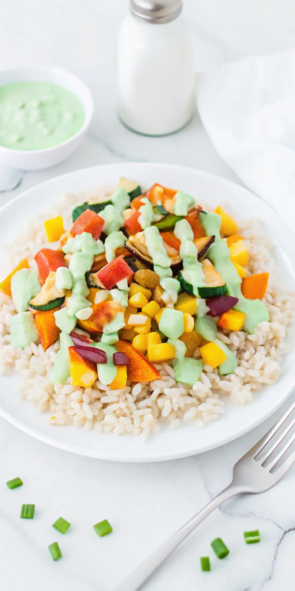 A close-up, slightly angled shot focusing on the textured surface of fluffy brown rice and the vibrant, roasted vegetables on a minimalist white plate. The creamy, pale green Green Onion Cashew Sauce is visibly drizzled, highlighting its smooth, thick consistency. A few chopped green onions are scattered for garnish. The background features a subtle wood accent and soft natural morning light on a marble countertop, emphasizing clean presentation and warm tones. NO HANDS.