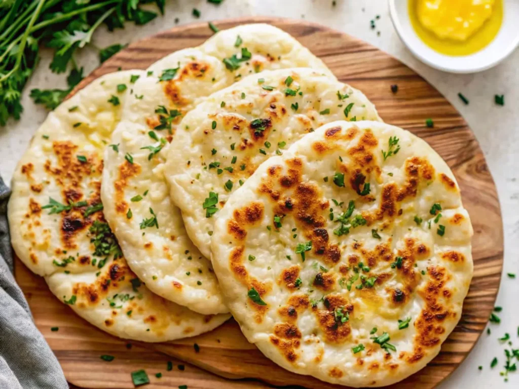 A hero shot of four golden-brown, fluffy cottage cheese flatbreads, generously drizzled with olive oil and sprinkled with fresh chopped parsley, arranged on a rustic wooden cutting board. The background features a marble countertop, a small ceramic bowl filled with fresh green herbs, and a small glass of olive oil, all bathed in soft natural morning light from an east window. Soft shadows, warm tones, clean and tidy presentation, no hands or people. (4:3 ratio)