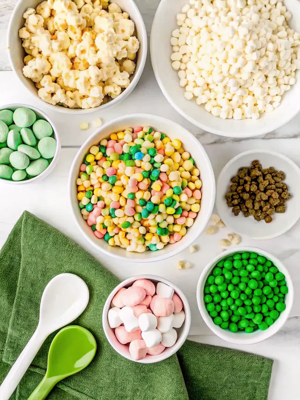 A minimalist arrangement of ingredients for 'Loaded Leprechaun Popcorn': a white ceramic bowl filled with unpopped popcorn kernels, a stack of colorful Lucky Charms marshmallows, a small dish of vibrant green candies, and a block of white chocolate and butter on a familiar wooden cutting board. All are neatly arranged on a white marble countertop under natural morning light from an east window. Fresh herbs in a small pot are subtly blurred in the background. The scene is clean and tidy. (3:4 ratio). NO HANDS OR PEOPLE.