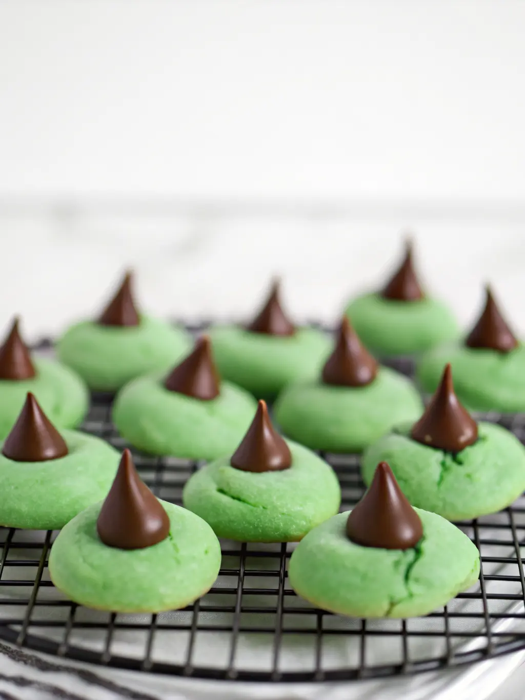 Ingredients laid out for Mint Chocolate Kiss Blossom Cookies: bowls of flour, sugar, and green dough, with unwrapped Hershey's Kisses and a bottle of peppermint extract. All arranged on a wooden cutting board against a marble countertop, under natural morning light, with fresh herbs subtly in the background. (3:4 ratio)