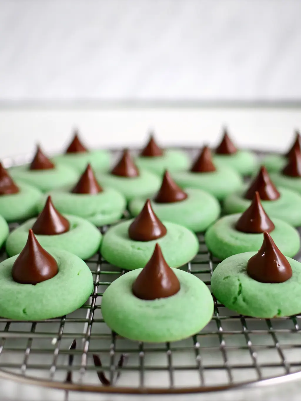 A moment in the process of making Mint Chocolate Kiss Blossom Cookies: green cookie dough balls, freshly rolled in granulated sugar, waiting on a parchment-lined baking sheet on a wooden cutting board. A single unwrapped Hershey's Kiss sits nearby. Natural morning light, soft shadows, warm tones, clean kitchen. (3:4 ratio)