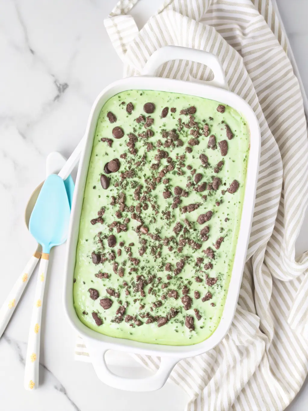 Overhead shot of ingredients for Shamrock Lasagna: packages of Oreo cookies, a block of cream cheese, a container of whipped topping, a bottle of mint extract, and green food coloring, all neatly arranged on a wooden cutting board on a clean marble countertop, with fresh herbs subtly in the background. Natural morning light, soft shadows, warm tones. No hands or people. (3:4 ratio)
