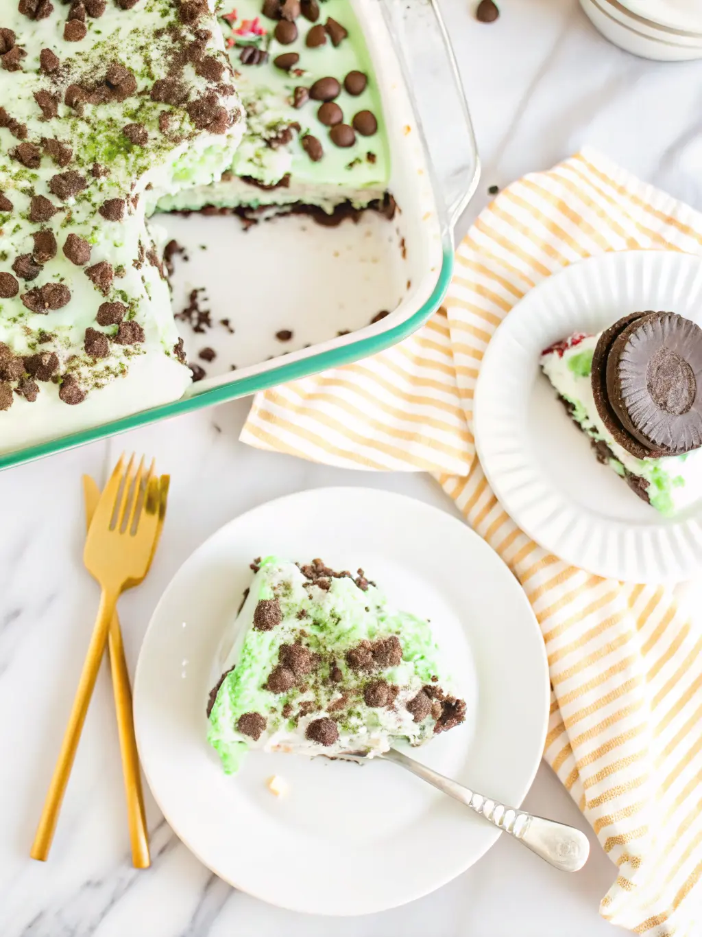 A close-up of the layering process for Shamrock Lasagna in a glass baking dish, showing the dark Oreo crust, the white cream cheese layer partially spread, and a spoonful of the vibrant green mint layer being gently added. The layers are distinct. Shot on a marble countertop with natural morning light, soft shadows, warm tones, with minimalist ceramic bowls in the soft background. No hands or people. (3:4 ratio)