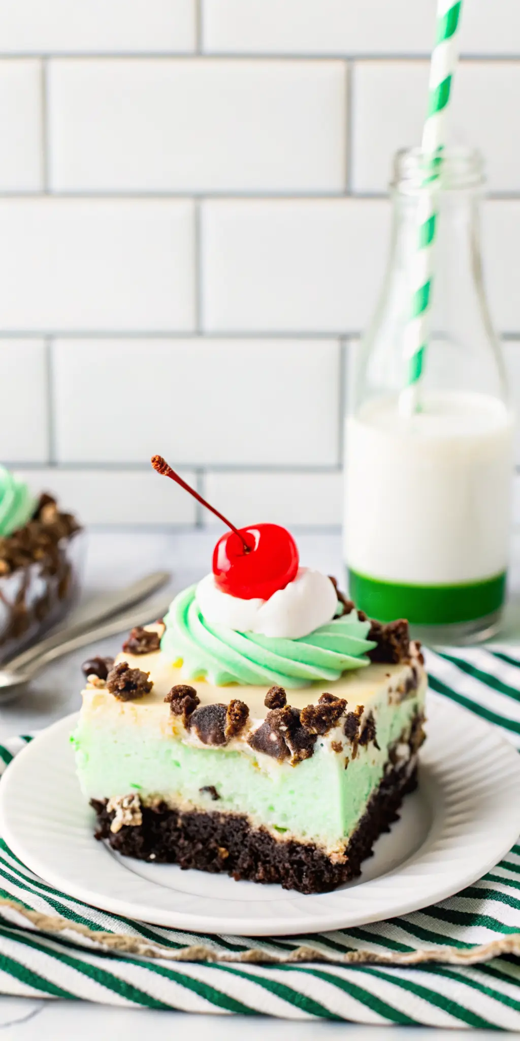 A close-up, slightly angled shot of a single slice of Shamrock Lasagna on a minimalist white plate, revealing the perfect, firm layers of Oreo crust, white cream cheese, and bright mint green. The top is generously adorned with whipped cream, crushed Oreos, and vivid green sprinkles, with a single maraschino cherry. The texture of the layers is clearly visible. A golden fork is nearby. Shot with natural morning light, soft shadows, warm tones, on a clean marble countertop. No hands or people. (3:4 ratio)