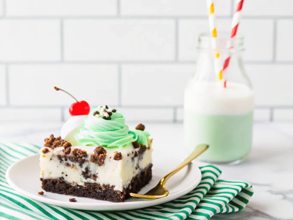 A generous slice of St. Patrick's Day Shamrock Lasagna dessert on a minimalist white plate, showcasing distinct layers of dark Oreo crust, light cream cheese, and vibrant mint green, topped with fluffy white whipped cream, crushed Oreos, green sprinkles, and a bright red maraschino cherry. A golden fork rests beside the plate on a yellow and white striped linen. A glass bottle of milk with a blue striped straw is in the soft focus background. Shot with natural morning light from an east window, soft shadows, warm tones, clean marble countertop with subtle wood accents in the distance. No hands or people. (4:3 ratio)