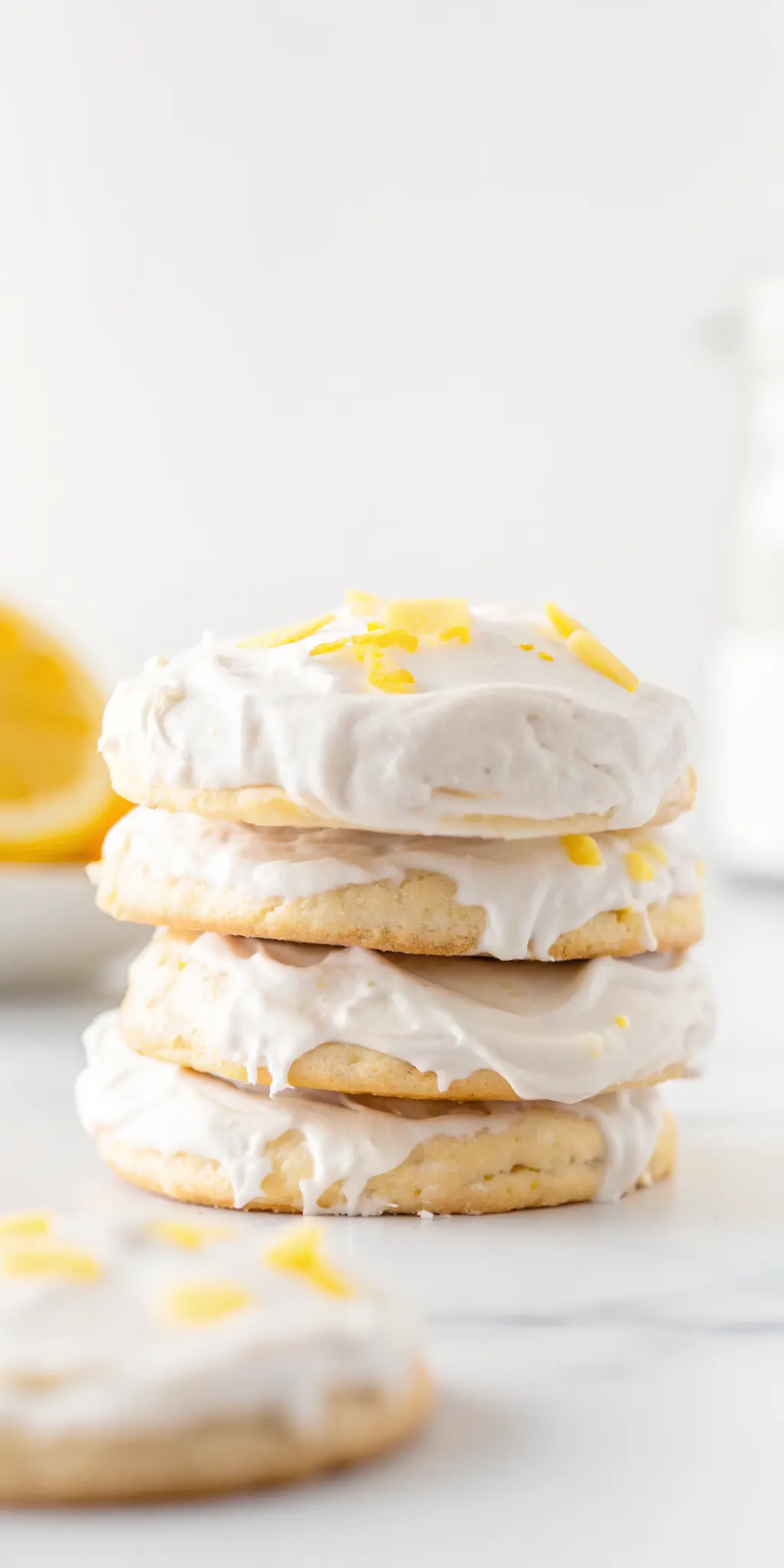 A soft-baked vegan lemon cookie, perfectly frosted with white lemon glaze and sprinkled with bright yellow lemon zest, resting on a minimalist white ceramic plate. The plate is on a white marble countertop, with soft natural morning light creating gentle shadows. Fresh herbs are blurred subtly in the background. Warm tones, clean and tidy presentation. 3:4 ratio. NO HANDS.