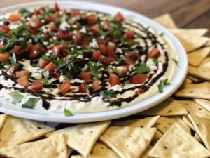 A vibrant, close-up shot of the Bruschetta Dip on a minimalist white plate, surrounded by golden pita chips. The dip features layers of creamy white base, a golden drizzle, and is topped generously with finely diced red Roma tomatoes, fresh green basil, and dark balsamic glaze. Shot on a marble countertop with soft natural morning light from an east window, casting gentle shadows. Warm tones, clean and tidy presentation. NO HANDS OR PEOPLE. (4:3 ratio)