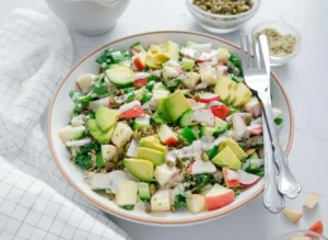 A vibrant, fresh Vegan Protein Kale Salad in a minimalist white bowl with a thin brown rim, overflowing with chopped kale, fluffy quinoa, diced cucumber, sliced radishes, vibrant red apple pieces, and perfectly fanned avocado slices. Creamy white dressing is drizzled over the top, with a small white ceramic pitcher of dressing nearby. A small clear glass bowl of pumpkin seeds and another with hemp seeds are visible in the background. A vintage-style silver fork and knife rest beside the main bowl. The scene is set on marble countertops with subtle wood accents, bathed in natural morning light from an east window, with soft shadows and warm tones. Fresh herbs are artfully blurred in the background, with a clean, tidy presentation. No hands or people. (4:3 ratio)