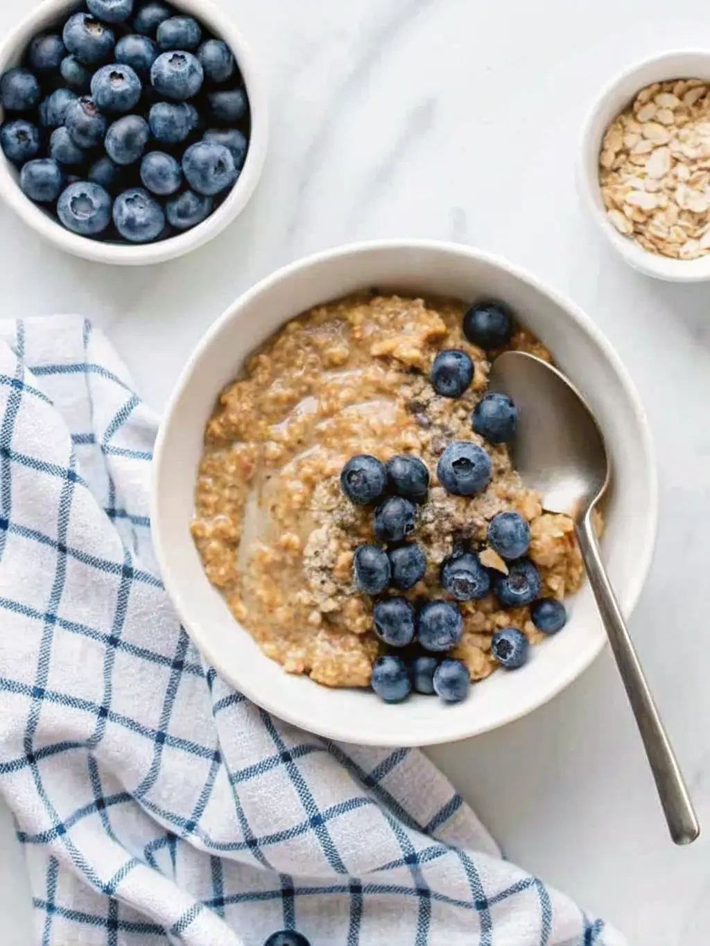 A collection of ingredients for a Vegan Quinoa Breakfast Bowl: dry quinoa in a small ceramic dish, fresh blueberries in a separate bowl, a jar of almond butter, a plant milk carton, cinnamon sticks, and vanilla extract. All arranged on a light marble countertop with the same wooden cutting board in the background. Natural morning light from an east window, soft shadows, warm tones. A small bunch of fresh basil (herbs) in the background. Clean and tidy presentation. NO HANDS OR PEOPLE.