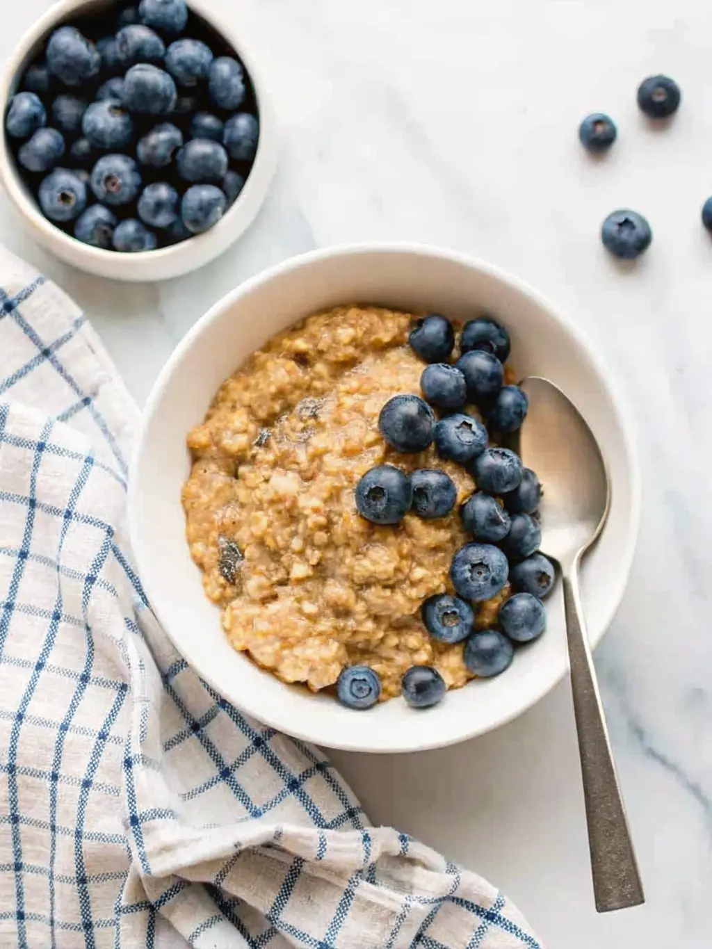 Close-up, vertical shot (3:4) focusing on the creamy texture of a Vegan Quinoa Breakfast Bowl in a white ceramic bowl, showcasing the fluffy quinoa, glistening blueberries, and the smooth swirl of almond butter, with visible hemp seeds. The bowl is on a light marble countertop, with a hint of the blue and white checkered towel. Natural morning light, soft shadows, warm tones. Fresh thyme sprigs (herbs) in the background. Clean and tidy presentation. NO HANDS OR PEOPLE.