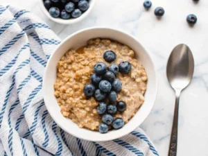 Hero shot, overhead view of a creamy Vegan Quinoa Breakfast Bowl in a minimalist white ceramic bowl, generously topped with vibrant fresh blueberries, a rich drizzle of almond butter, and a sprinkle of hemp seeds. A silver spoon rests beside the bowl on a light marble countertop. A blue and white checkered kitchen towel is neatly folded next to the bowl. Natural morning light from an east window casts soft shadows. A small bunch of fresh mint leaves (herbs) in a glass jar in the soft background. The same wooden cutting board is slightly visible in the background. Warm tones, clean and tidy presentation. NO HANDS OR PEOPLE.