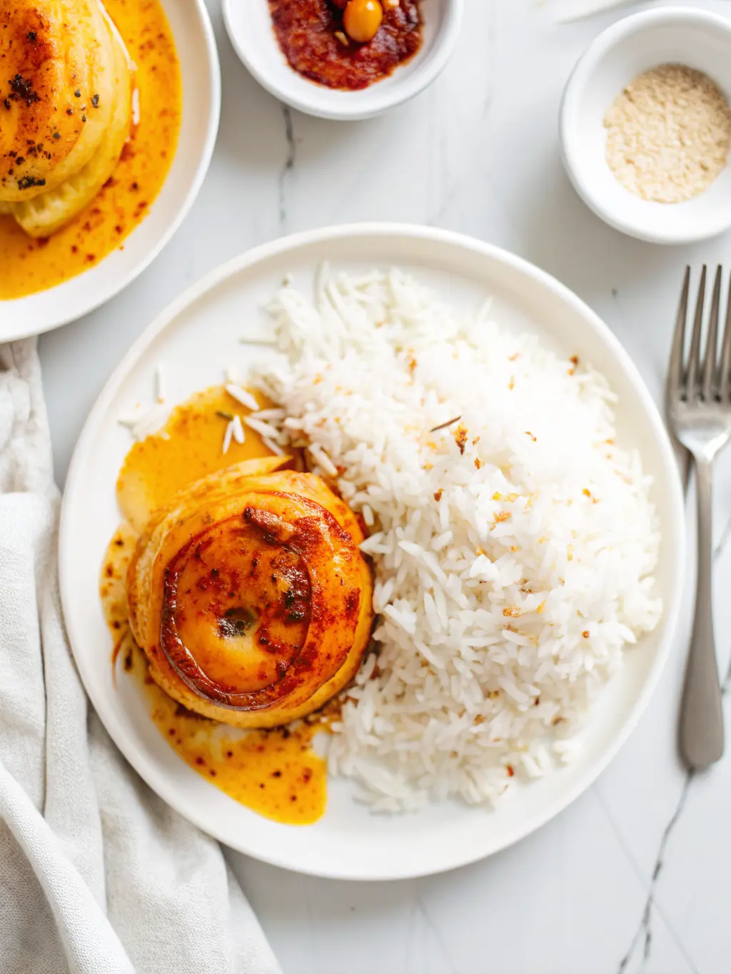 A close-up, slightly overhead shot of a single, finished Onion Boil on a minimalist white plate. The onion is tender and translucent, generously coated in vibrant red and orange spices, and bathed in a glistening pool of savory, orange-red oil/sauce. A subtle cut on top reveals the soft interior. The plate rests on a marble countertop, with warm tones and soft shadows. Fresh herbs are blurred in the background. No hands or people. (3:4 ratio)