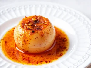 A single, perfectly cooked whole Onion Boil, richly seasoned with vibrant red and orange spices, glistening with a generous pool of orange-red, savory oil/sauce, centered on a minimalist white plate with a subtle rim design. The scene is bathed in natural morning light from an east window, sitting on a marble countertop with a subtle wood accent in the soft-focused background, clean and tidy presentation, soft shadows and warm tones. No hands or people.