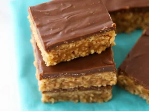 A hero shot (4:3 ratio) of a stack of three no bake Scotcheroo bars, showcasing the distinct layers of crispy peanut butter rice cereal, a subtle caramel-like middle, and a smooth dark chocolate topping. The bars are resting on a teal/turquoise napkin on a clean marble countertop, with soft natural morning light from an east window casting gentle shadows. Warm tones, clean and tidy presentation with a blurred background.