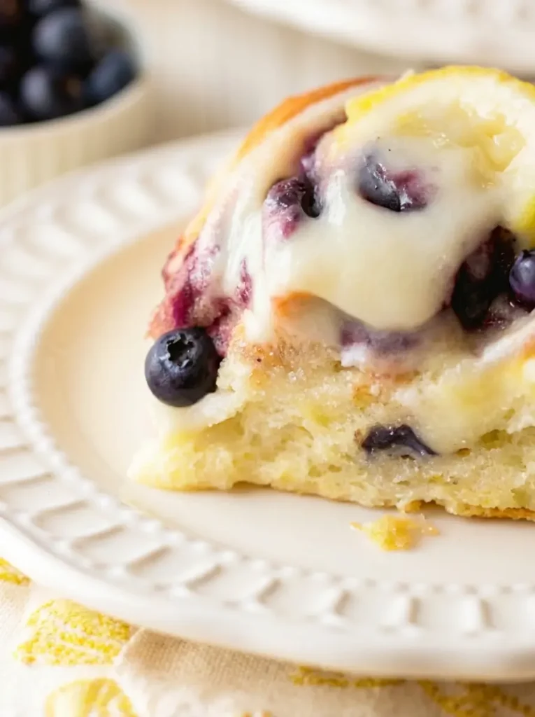Blueberry Lemon Sweet Rolls A shot of the preparation process on a marble countertop with wood accents. A sheet of yeast dough is rolled out flat. Fresh, vibrant blueberries are being scattered over the dough. A wooden rolling pin and a zester with a fresh lemon are visible on the side. The lighting is bright and airy. 3:4 aspect ratio.