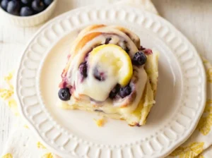 A high-angle hero shot of a single Blueberry Lemon Sweet Roll on a decorative white ceramic plate with subtle scalloped edges. The roll is golden brown, swirled with dark cooked blueberries, and heavily glazed with white icing. A small, fresh lemon wedge sits perfectly on top. The background is a blurred kitchen scene with a marble countertop, a wooden cutting board, and a bowl of fresh blueberries. Natural morning light from the east creates soft shadows. 4:3 aspect ratio.