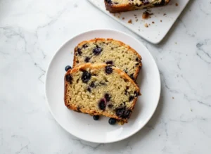 A high-angle 4:3 hero shot of two thick slices of blueberry muffin bread stacked slightly offset on a minimalist white ceramic plate. The bread has a golden-brown, sugary crust and a moist, yellowish crumb generously studded with dark purple baked blueberries. Several fresh blueberries are scattered casually on the white plate. The setting is a marble countertop with soft morning light streaming from the right, casting gentle shadows. A wooden cutting board and a blurred pot of fresh herbs are visible in the background, maintaining the warm, clean kitchen aesthetic.