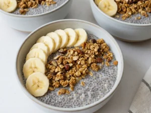 A hero shot of three glass mason jars filled with creamy overnight oats, sitting on a marble countertop with warm wood accents. The center jar is topped with perfectly arranged banana slices in a shingle pattern. Visible black chia seeds speckle the creamy beige oat mixture. Soft morning light streams from the east, casting gentle shadows. A white linen napkin is casually draped to the left. No hands or people.