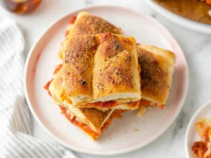 A high-angle close-up shot of a stack of two pizza sliders on a minimalist white plate with a raised rim. The sliders feature golden-brown Hawaiian roll tops glistening with garlic butter and herbs. Layers of red pepperoni, rich tomato sauce, and thick melted white mozzarella cheese are visible and oozing from the sides. The plate sits on a grey marble coaster. In the background, soft focus white subway tiles and a grey linen napkin. Natural morning light from the east creates soft shadows. 4:3 aspect ratio.
