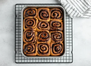 A high-angle, straight-down hero shot of a rectangular metal baking pan filled with 6 visible chocolate cinnamon rolls. The rolls have a golden-brown crust and a distinct dark chocolate spiral filling with visible texture of chocolate crumbs. The pan sits on a marble countertop with soft morning light casting gentle shadows from the east. A wooden cutting board is partially visible in the periphery.