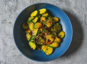 A vibrant green Easy Asian Cucumber Salad, made with smashed cucumber slices, glistening with a reddish-brown savory-spicy dressing, generously topped with white sesame seeds and red chili flakes, presented in a minimalist white ceramic bowl on a marble countertop with subtle wood accents. Natural morning light from an east window casts soft shadows. Fresh herbs are visible in the soft background, with warm tones and a clean, tidy presentation. No hands or people.