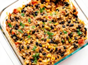 A top-down shot of a golden-brown and bubbly Easy Chicken Burrito Casserole, freshly baked in a clear glass baking dish, generously topped with shredded cheese, black beans, corn, red bell peppers, and fresh green cilantro and sliced green onions. The casserole is placed on a light marble countertop with a subtle wood accent visible, bathed in natural morning light from an east window, casting soft shadows. The overall presentation is clean and tidy, with warm tones, no hands or people. (4:3 ratio)