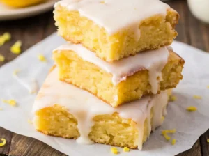 A stack of two bright yellow lemon brownie squares, one with a bite taken out revealing the moist, tender crumb texture, generously topped with a thick, smooth white lemon glaze and sprinkled with fresh lemon zest, resting on white parchment paper. The setting is a clean marble countertop with soft natural morning light from an east window, blurred wooden accents, and a hint of fresh lemons and green herbs in the background. Warm tones, soft shadows, clean and tidy, 4:3 ratio.