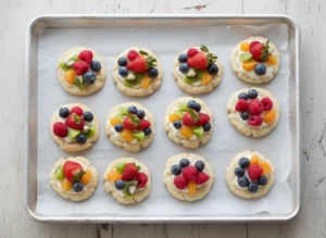 A hero shot of several vibrant Fruit Pizza Cookies arranged artfully on a minimalist white ceramic platter, showcasing their soft sugar cookie base, creamy white frosting, and colorful toppings of fresh blueberries, raspberries, diced kiwi, sliced strawberries, and mandarin orange segments. The scene is bathed in natural morning light from an east window, casting soft shadows on marble countertops with subtle wood accents in the background. Fresh herbs are visible in a small ceramic bowl.