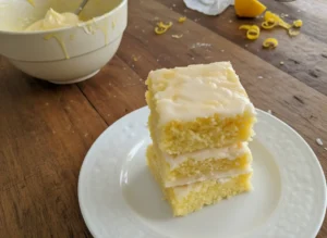 A stack of three light yellow lemon brownies with a white glaze, on a green ceramic plate, resting on a white linen napkin. The scene is set on a warm-toned wooden countertop in a bright kitchen with natural morning light coming from an east window, a silver stand mixer subtly blurred in the background, fresh whole lemons in a minimalist white ceramic bowl, and a white subway tile backsplash. Soft shadows, warm tones, clean and tidy presentation, no hands or people. (4:3 ratio)