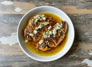 A high-angle, close-up hero shot of golden-brown pan-seared chicken breasts sitting in a pool of golden lemon-olive oil sauce. The chicken is generously topped with chunky white crumbled feta cheese and vibrant green chopped fresh dill and parsley. The food is plated on a minimalist white ceramic platter sitting on a marble countertop with warm wood accents. Soft, natural morning light from the east creates gentle shadows. No hands or people.