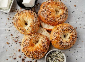 A close-up, inviting hero shot of several golden brown bagels, perfectly round and heavily coated in everything bagel seasoning (white sesame, black sesame, poppy seeds, dried garlic flakes, dried onion flakes), arranged on a rustic wooden cutting board on a clean white marble countertop. Strong natural morning light from an east window casts soft, warm shadows. The background is subtly blurred, showing a minimalist ceramic bowl with fresh chives.