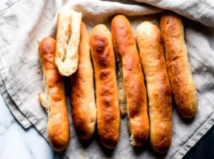 A close-up, top-down shot of several golden-brown, elongated homemade soft breadsticks artfully arranged on a clean marble countertop, with one breadstick broken in half revealing its soft, fluffy white interior texture. A red and white checkered kitchen towel is casually draped in the soft background. The scene is bathed in natural morning light from an east window, with subtle soft shadows and warm tones, embodying a clean and tidy presentation, no hands or people.