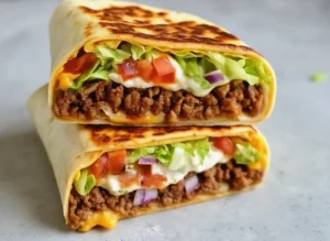 A high-angle hero shot of a stack of three sliced Keto Crunchwraps on a minimalist white ceramic plate. The cut sides are facing the camera, revealing distinct layers of browned ground beef, melted orange cheddar, a thin crispy interior line, white sour cream, green lettuce, and red tomatoes. The wraps are golden-brown and toasted. The setting is a bright kitchen with marble countertops, soft morning light coming from the right, and a blurry background of fresh cilantro and a wooden cutting board.