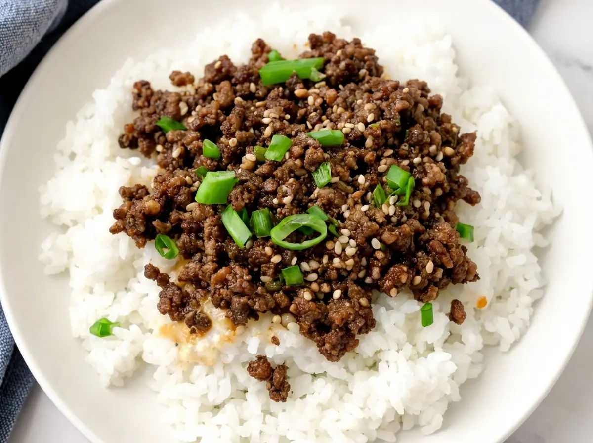 A vibrant Korean Ground Beef Bowl, piled high with glossy, seasoned ground beef, sprinkled with white sesame seeds and fresh green onion slices, resting on a bed of fluffy white rice in a minimalist white ceramic bowl. Wooden chopsticks are laid beside the bowl. The scene is set on a light grey marble countertop with a soft grey linen cloth draped nearby. Natural morning light from an east window casts soft shadows. Fresh green herbs are subtly blurred in the background, near a wooden cutting board, creating a clean, warm, and inviting atmosphere with warm tones. 4:3 ratio, no hands.