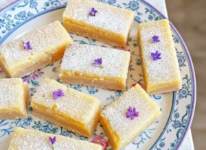 A close-up, top-down view of several golden rectangular Lavender Lemon Bars, dusted generously with powdered sugar and sprinkled with tiny purple culinary lavender flecks, arranged elegantly on a minimalist white ceramic plate with a subtle blue and purple floral pattern. The plate sits on a marble countertop, bathed in soft natural morning light from an east window, casting gentle shadows. A hint of a wooden cutting board is in the background, along with a blurred ceramic bowl. The scene is clean and tidy, with warm tones. (4:3 ratio)