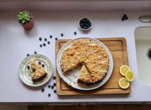 A whole Lemon Blueberry Ricotta Cake with a slice removed, revealing the moist, blueberry-studded interior and a golden streusel topping, drizzled with a white lemon glaze. It's perfectly presented on a minimalist white plate, resting on a rustic wooden cutting board on a light marble countertop. Scattered fresh blueberries and thin lemon slices adorn the surface, with natural morning light streaming from an east window, casting soft shadows. The background shows a tidy kitchen with subtle wood accents and fresh herbs in ceramic pots, all bathed in warm tones. (4:3 ratio)