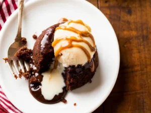 A close-up, overhead shot of a single dark chocolate lava cake on a minimalist white plate, partially eaten with a silver spoon revealing a rich, molten peanut butter and chocolate core. A generous scoop of creamy vanilla ice cream sits on top, adorned with thin drizzles of chocolate syrup and warm peanut butter sauce. The scene is bathed in natural morning light from an east window, casting soft shadows on marble countertops with subtle wood accents in the background. Fresh herbs are blurred in the far background, and a red and white checkered napkin is visible. The presentation is clean, tidy, and exudes warm tones. (4:3 aspect ratio)