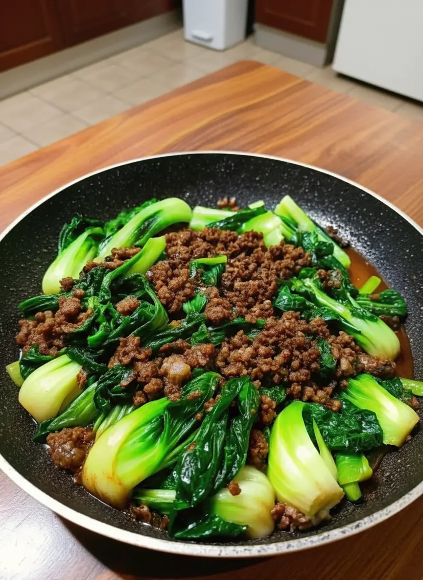 Action shot of the cooking process in a dark non-stick skillet. The ground beef is deeply browned and sizzling, pushed slightly to the side as bright green bok choy is being added to the pan. Steam rising softly, warm tones, no hands visible.