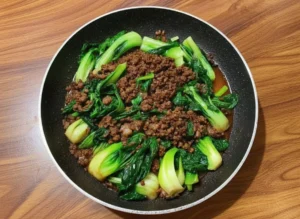A high-angle hero shot of a dark skillet resting on a marble countertop with wood accents. The skillet contains Stir-Fried Bok Choy With Beef featuring browned crumbled ground beef and bright green baby bok choy halves coated in a glossy dark brown sauce. Chopped green onions are scattered on top. Natural morning light from the east creates soft shadows. A wooden cutting board and fresh herbs are visible in the background.
