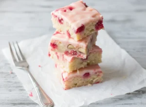 A stack of four square Strawberry Lemon Blondies on white parchment paper, centered on a light grey wooden cutting board with ornate silver forks peeking into the foreground. The blondies are pale golden with visible red strawberry chunks and a soft, light pink, slightly swirled lemon frosting. The scene is bathed in natural morning light from an east window, with soft shadows, warm tones, and a clean, tidy minimalist white plate visible out of focus in the background. No hands or people. (4:3 ratio)