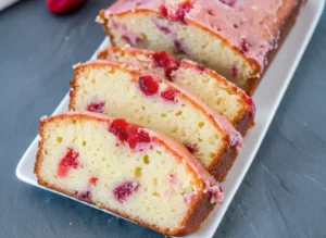 A perfectly glazed Strawberry Pound Cake loaf, partly sliced, showcasing the moist light yellow crumb and visible red strawberry pieces. The slices are arranged on a wooden cutting board on a marble countertop. Fresh whole strawberries are scattered nearby on a minimalist white plate. The scene is bathed in natural morning light, with soft shadows and warm tones, a clean and tidy presentation, shot at 4:3 ratio. No hands or people.