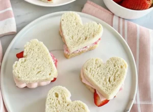 A beautifully arranged minimalist white plate filled with delicate heart-shaped strawberry tea sandwiches, some closed and some open-faced showcasing sliced fresh strawberries and creamy pink filling. A white ceramic bowl overflowing with whole ripe strawberries is blurred in the background. The scene is bathed in natural morning light, casting soft shadows on a light marble countertop with a subtle wood accent visible. Clean and tidy presentation, warm tones. (4:3 ratio)